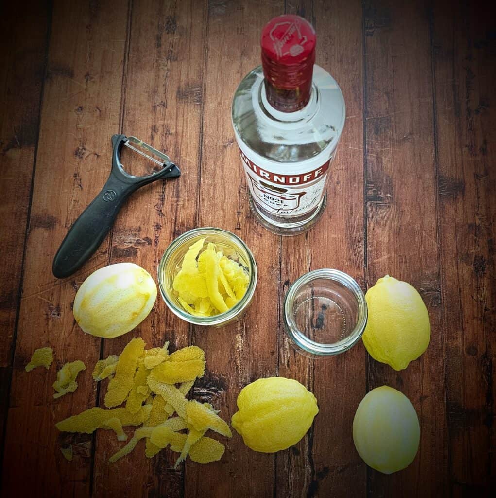 how to make lemon extract - how to make an extract - Fresh lemons, lemon zest, a vegetable peeler, two glass mason jars, and a bottle of vodka arranged on a wooden table for making homemade lemon extract.